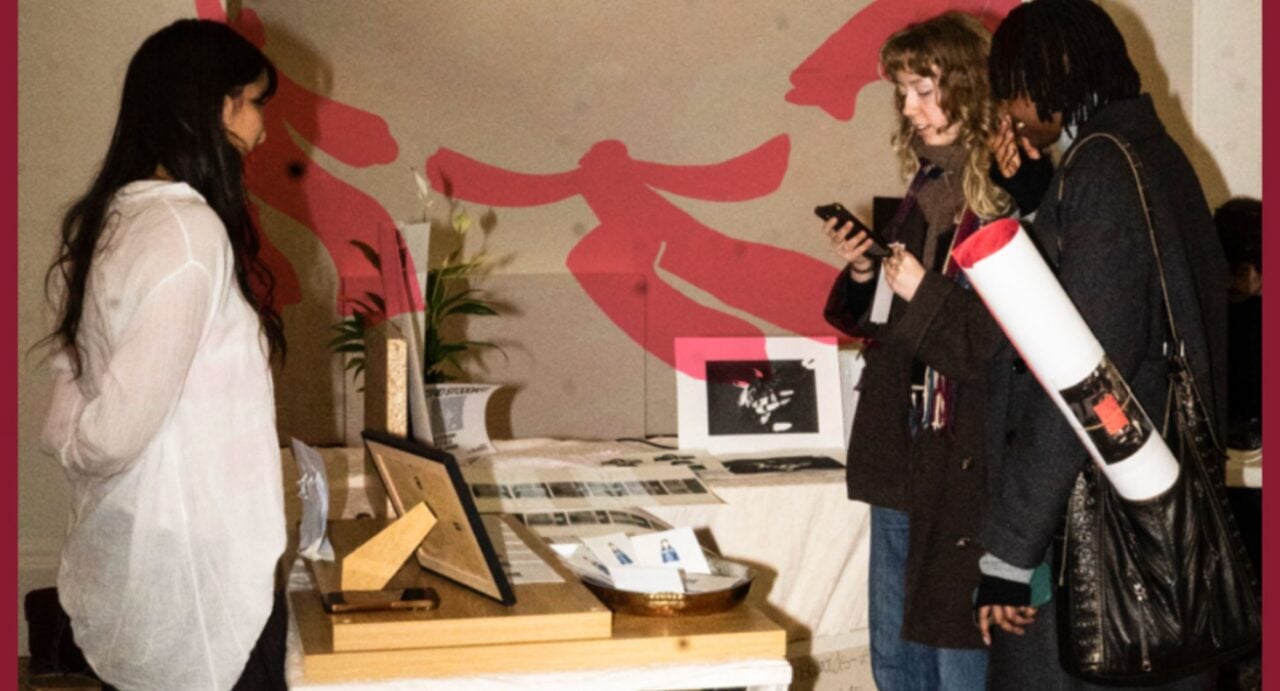 2 people look at a table and stand, on which are displayed prints and paintings.