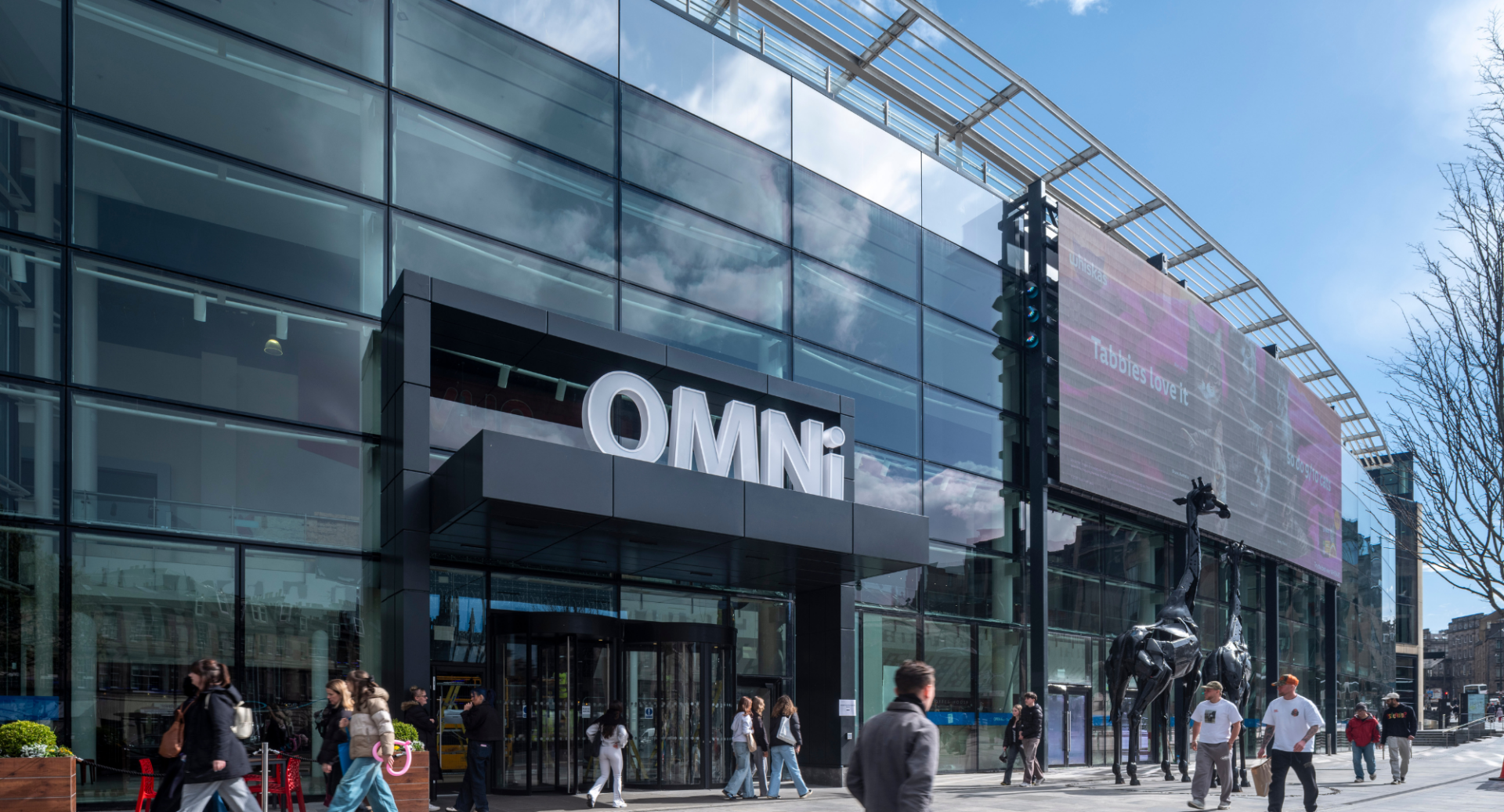 Glass exterior of a large shopping centre, with people passing by.