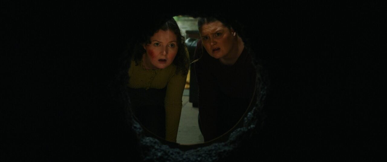 2 women looking through a hold in a wall.