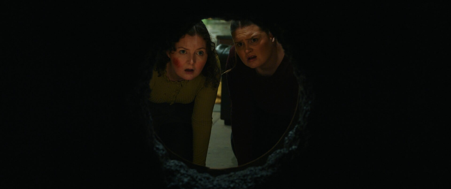 2 women looking through a hold in a wall.