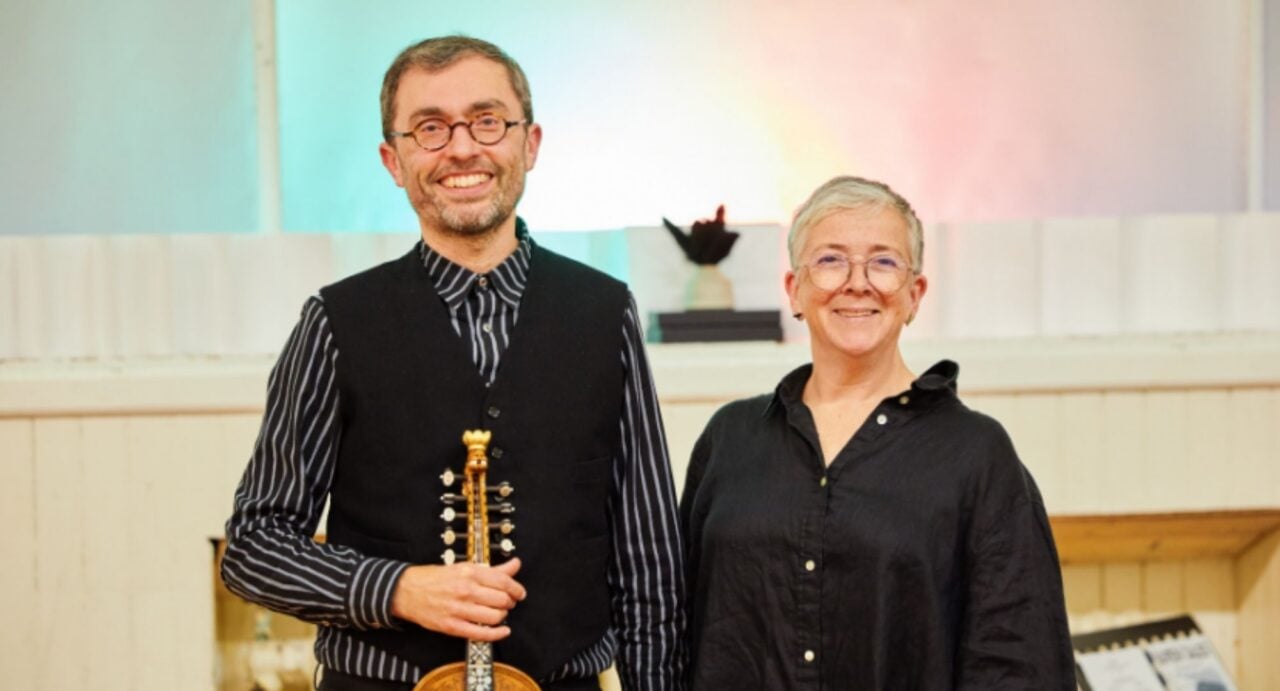 A man and a woman, both wearing black. The man is holding a musical instrument.