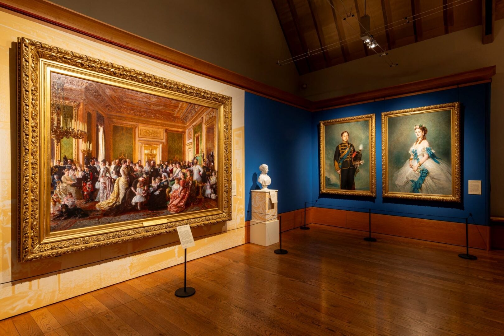 A painting of a large group of people on one wall, and on the adjacent wall, 2 full-length portraits.