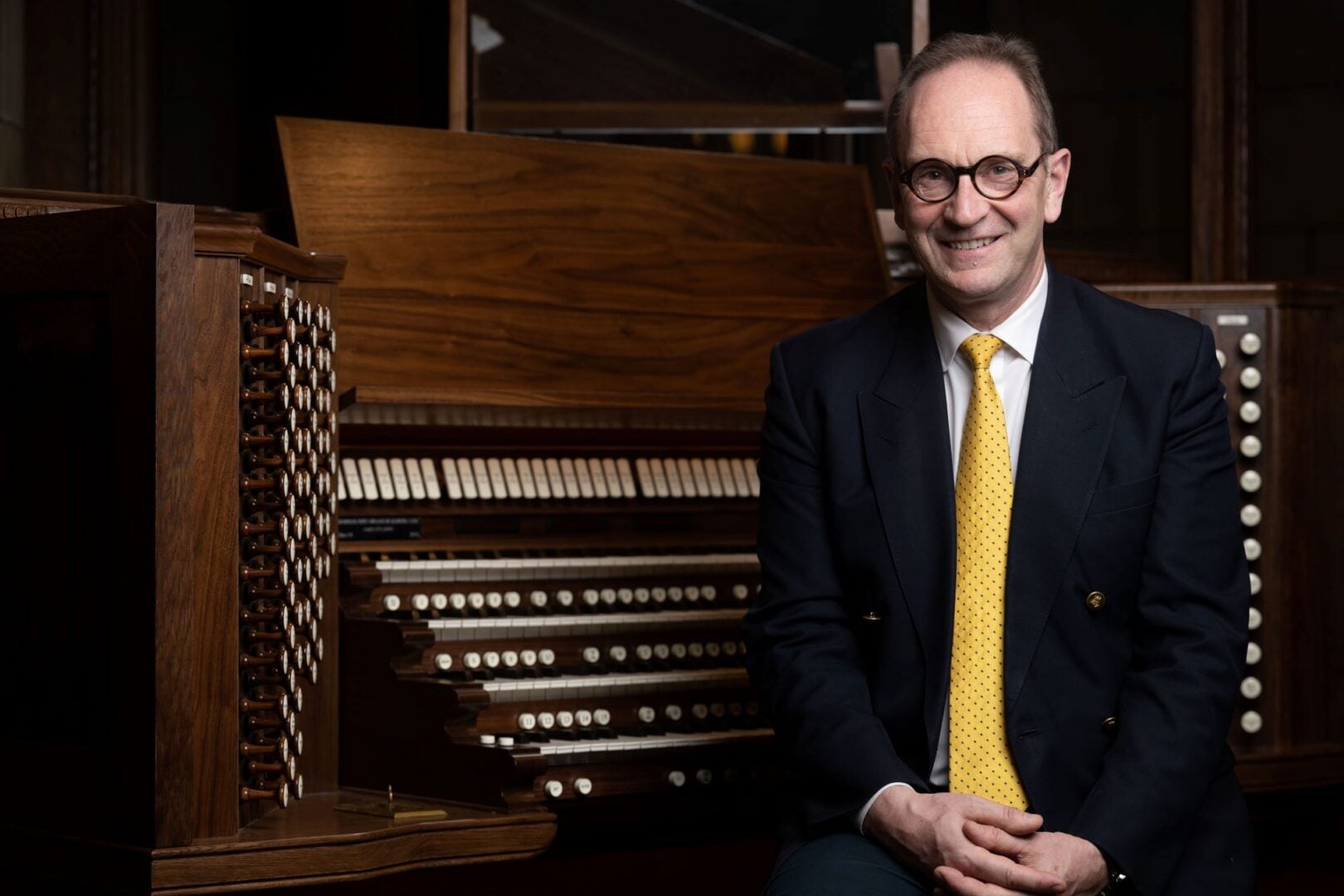A man wearing a dark suit and a yellow tie sitting at a large musical organ.