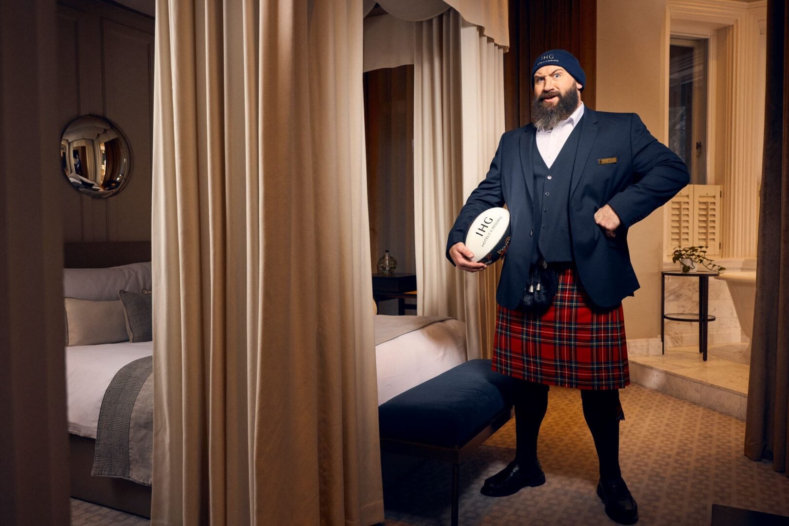 Man wearing a kilt and holding a rugby ball, standing in a hotel bedroom.