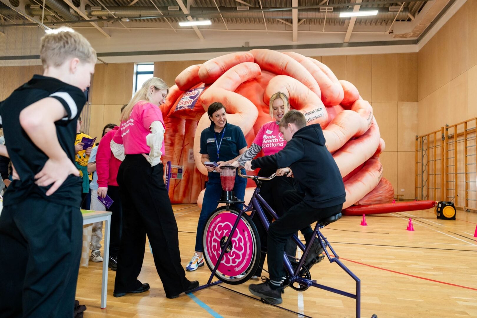 A young boy on a bike, surrounded by people looking on, and a large pink inflatable brain in the background.