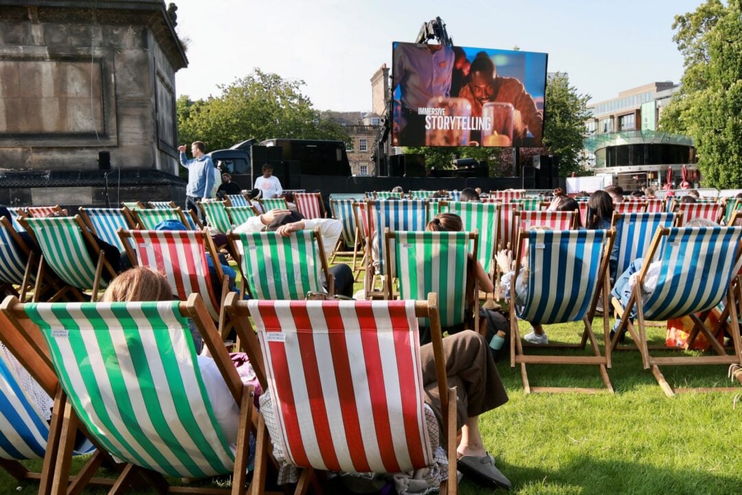 People sitting outdoors on deckchairs, looking at a large cinema screen.