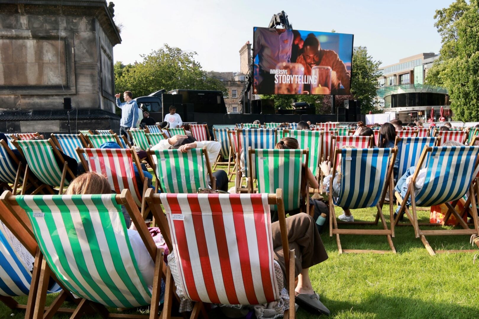 People sitting outdoors on deckchairs, looking at a large cinema screen.