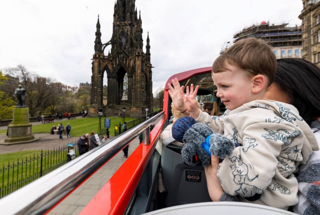 A young child on the red CitySightseeing Edinburgh Tour, waving to passers by, with Scott Monument in the background.,© Edinburgh Bus Tours