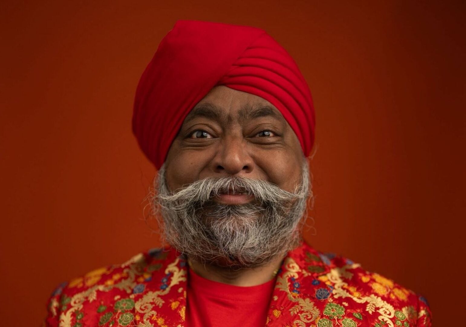 Head and shoulders image of a man wearing a red turban, and red and gold jacket.