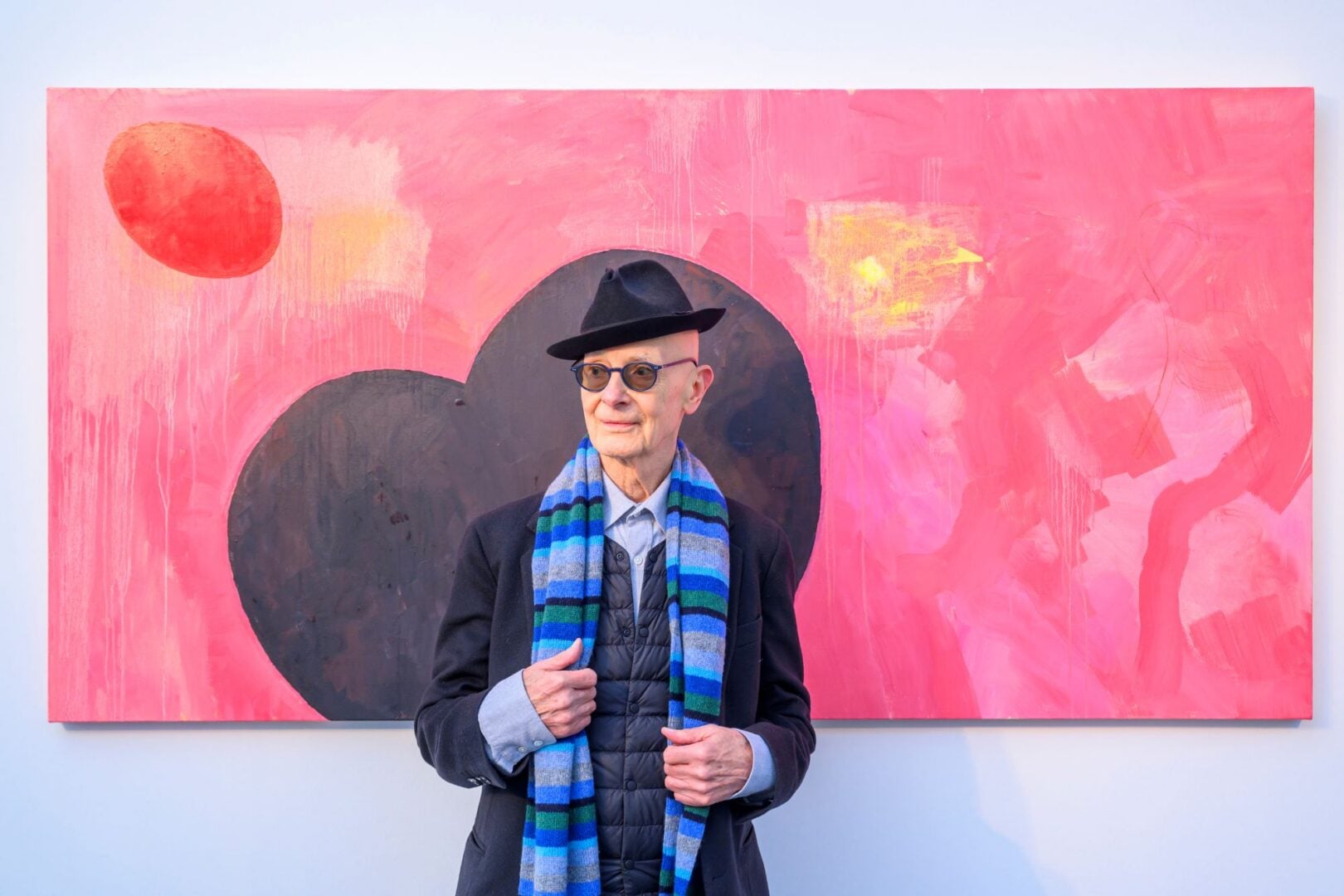 A man standing in front of a painting, in shades of pink, red, and black.
