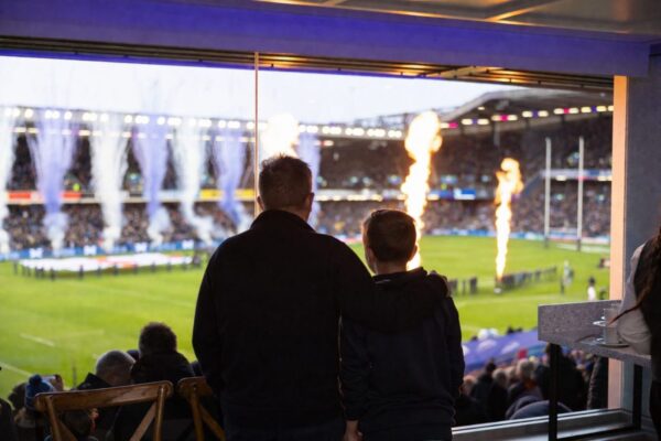 A man standing with his arm round a young boy, watching a game of rugby.