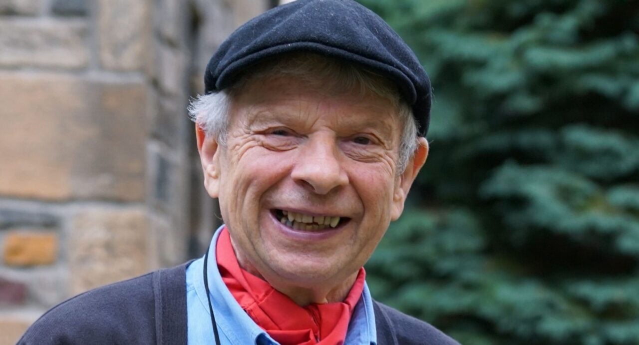 A smiling elderly man, wearing a blue cap and a red scarf.
