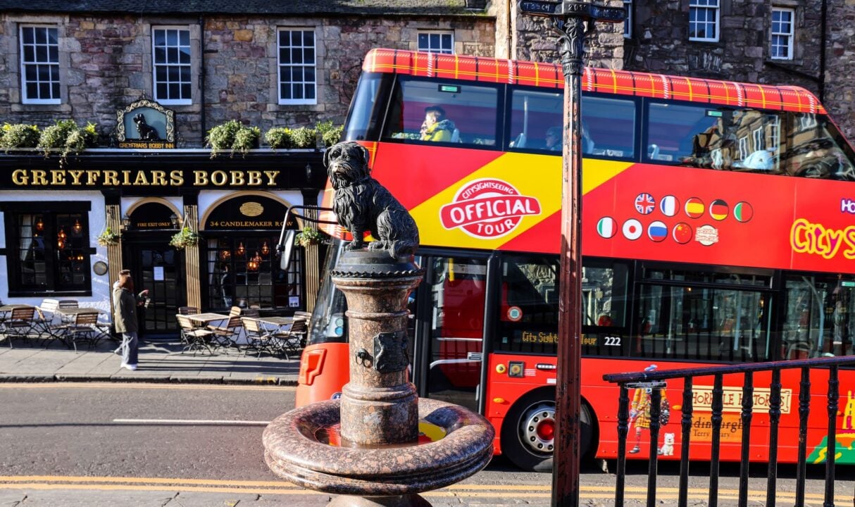 A red open-top tour bus going past a small bronze statue of a dog.