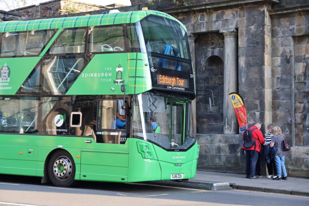 A green tour bus, with 2 people outside buying tickets from a ticket seller.