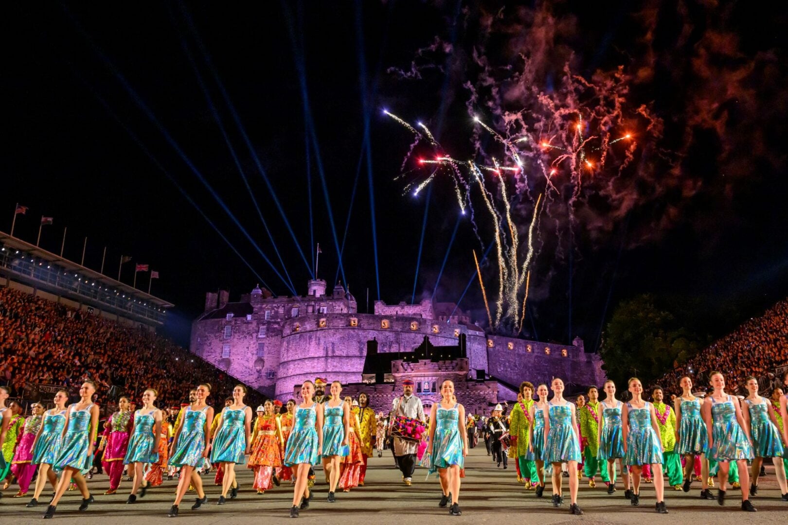 Rows of dancers performing in front of a castle at night-time, with fireworks going off in the sky.