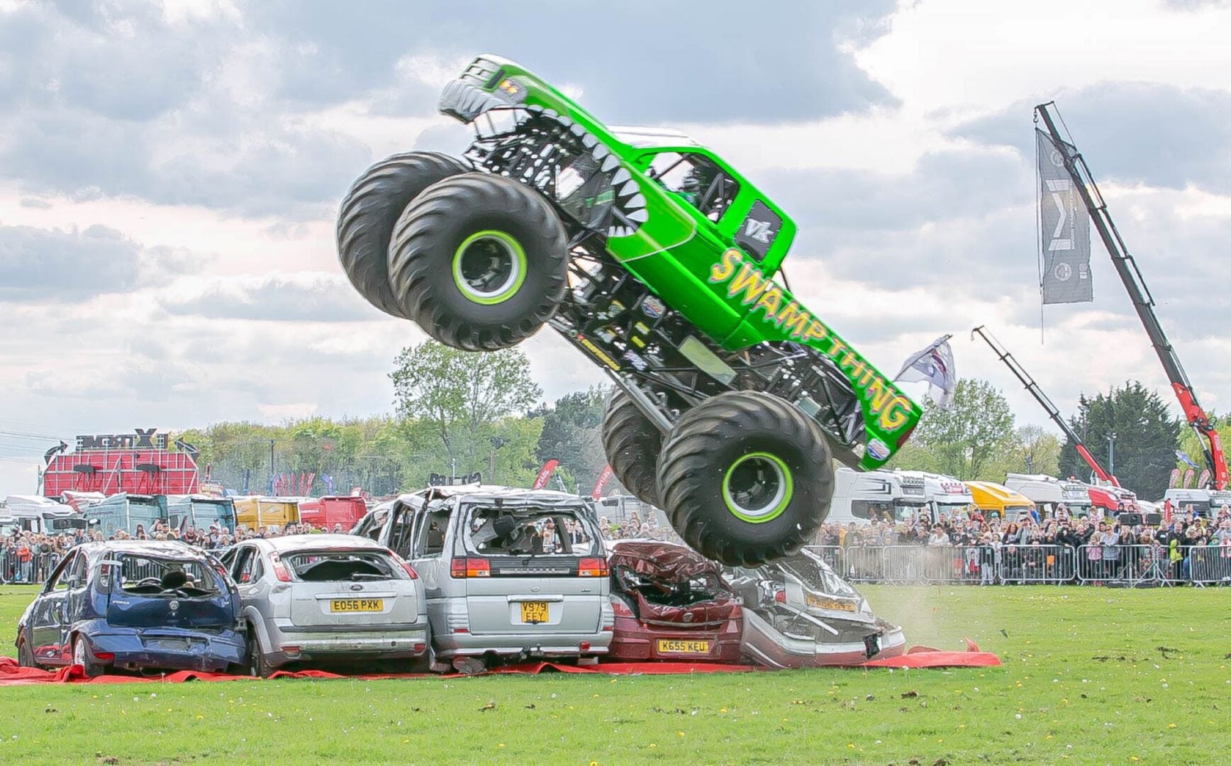 A large green truck riding over a row of 5 crushed cars.