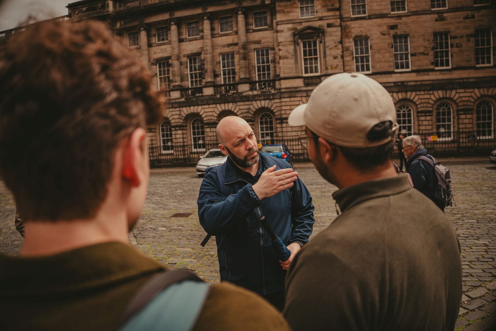 A man, with his back to a grand stone building, talking to a group of people.
