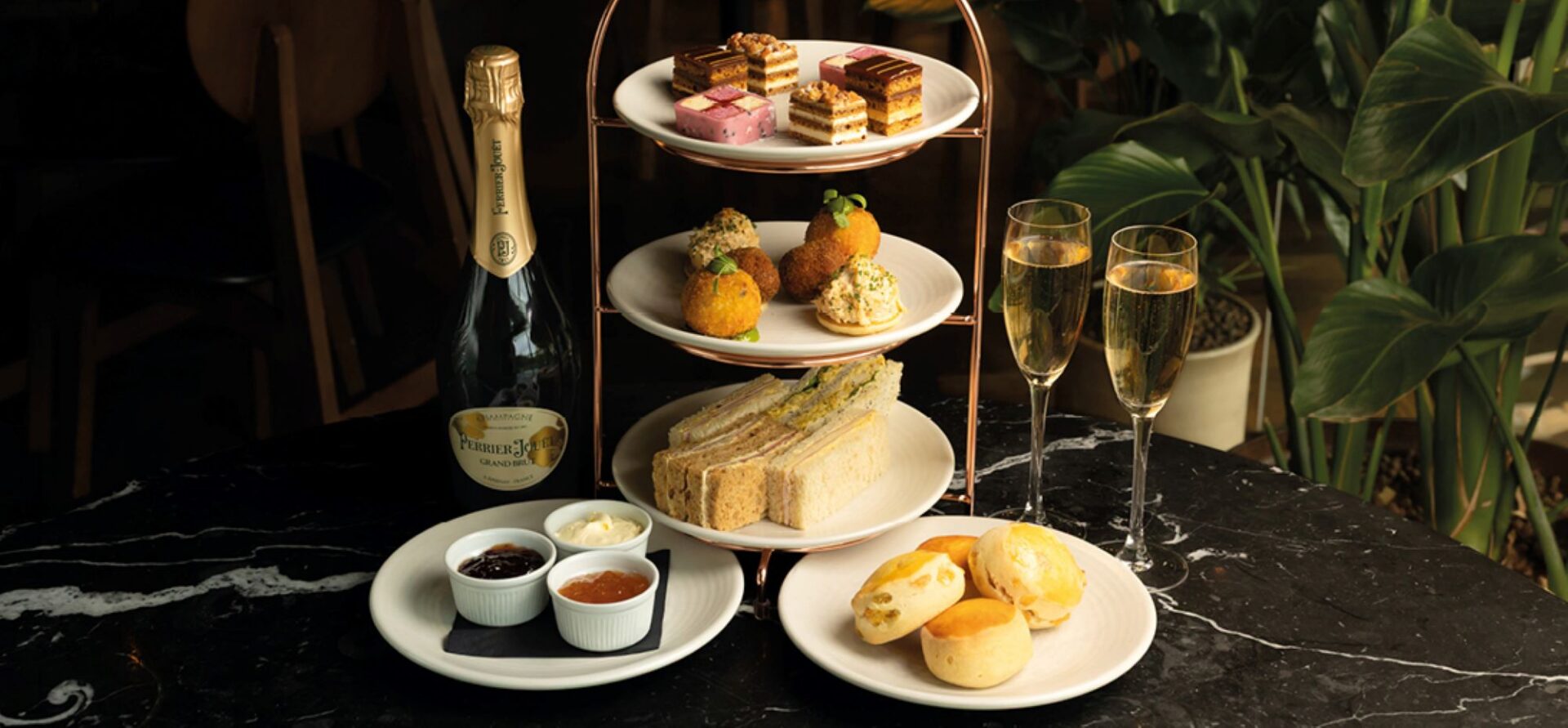 A 3-tier tea stand, with sandwiches, savoury bites and cakes, alongside plates of scones and jam, a bottle of champagne and 2 filled glasses.