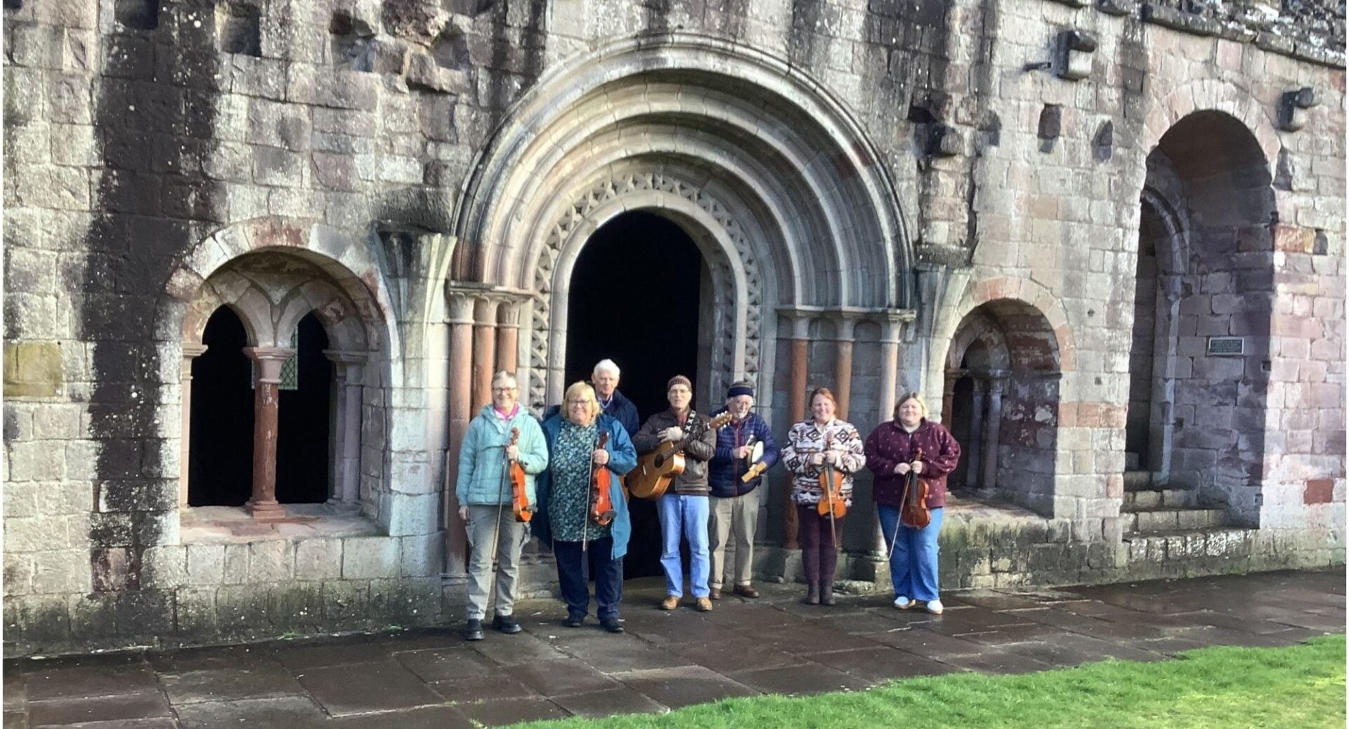 A group of people, holding instruments, standing at entrance of an old stone building.