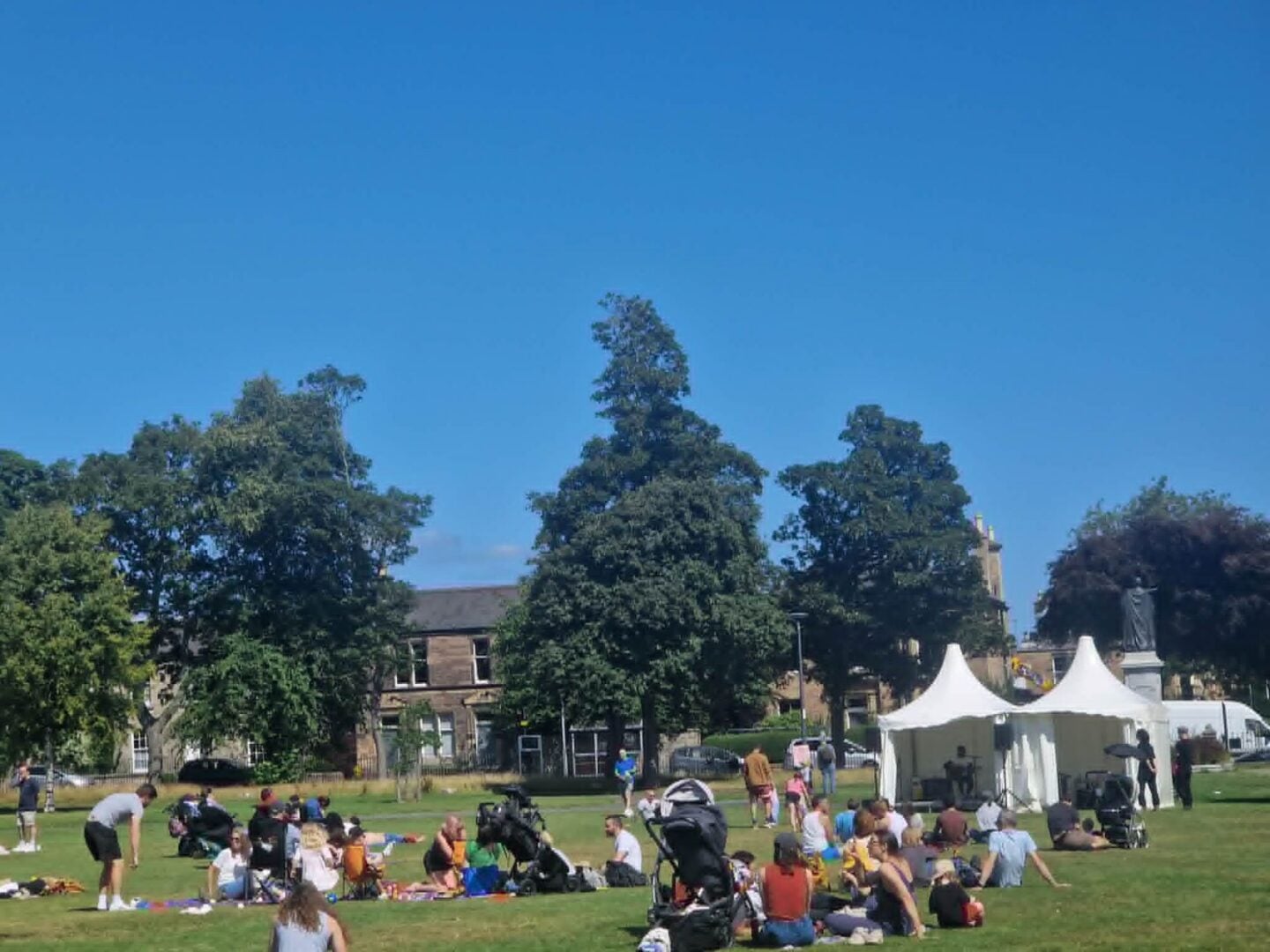 A sunny park, filled with people and a small white tent.