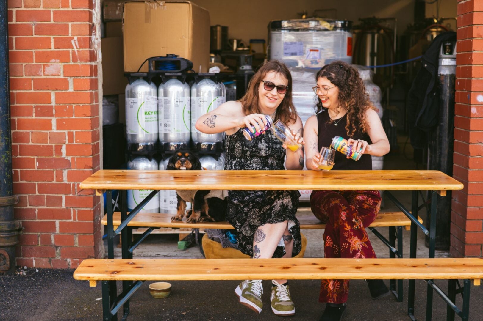 2 people seated at a table and bench, each pouring a glass of beer from a can.