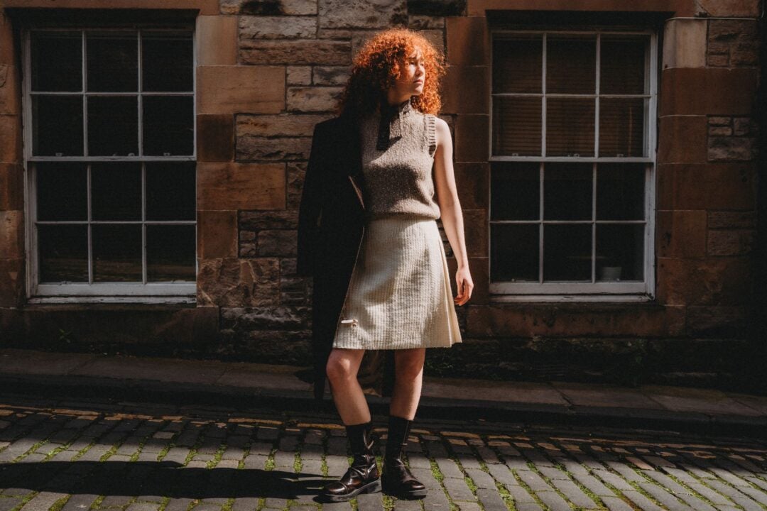 A woman with red hair wearing a beige wool top and kilt, standing on a cobbled street.