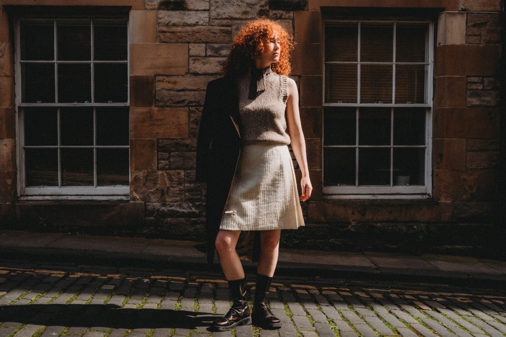 A woman with red hair wearing a beige wool top and kilt, standing on a cobbled street.