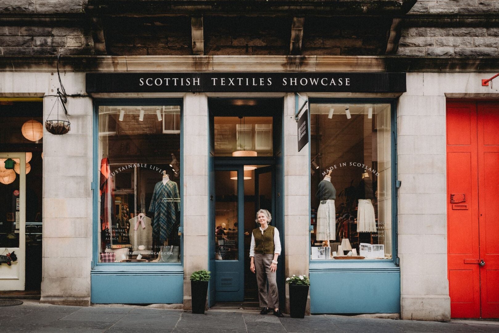 A woman standing outside a shop, which has displays of kilts and woollen clothes in the windows.