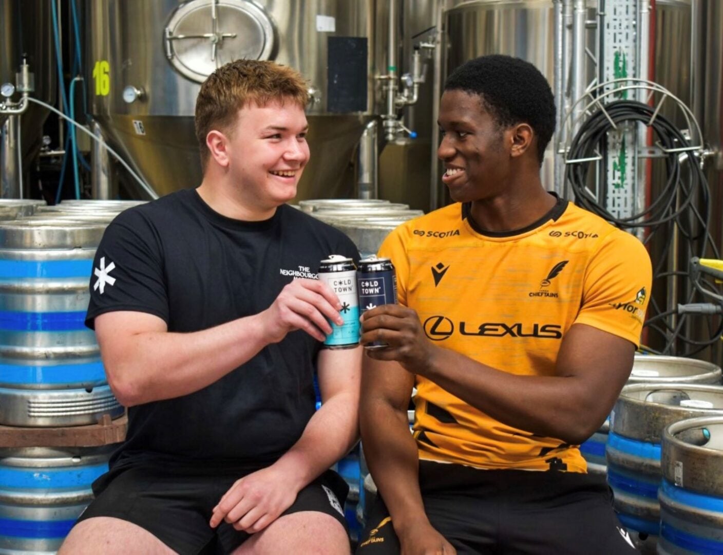 2 men, both dressed in sports clothing, saying cheers with a can of beer.