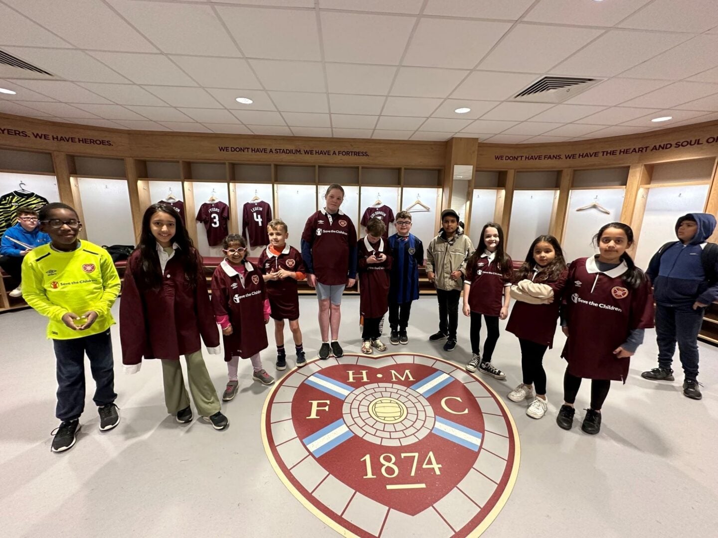 A group of young children, some wearing burgundy coloured football tops, in a stadium dressing room.