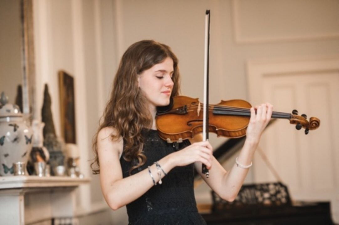 A woman playing a violin in a room with white panelled walls and fireplace.