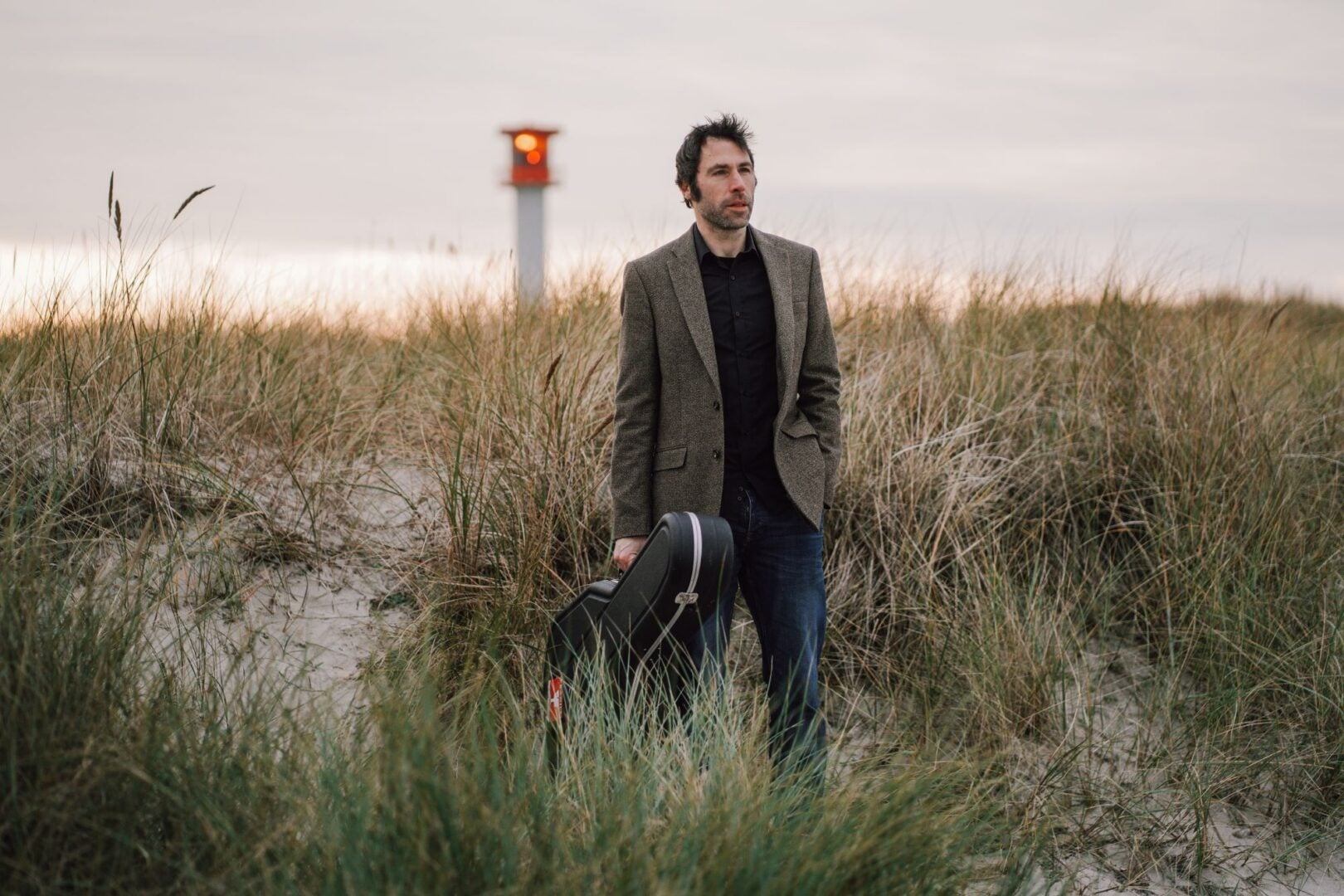 A man, holding a large musical instrument case, wandering through sandy dunes.