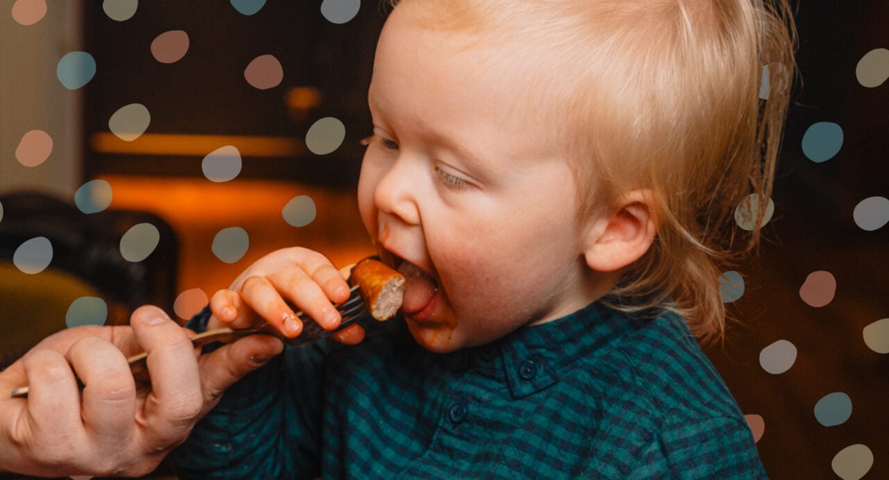 A young child eating a sausage from a fork.