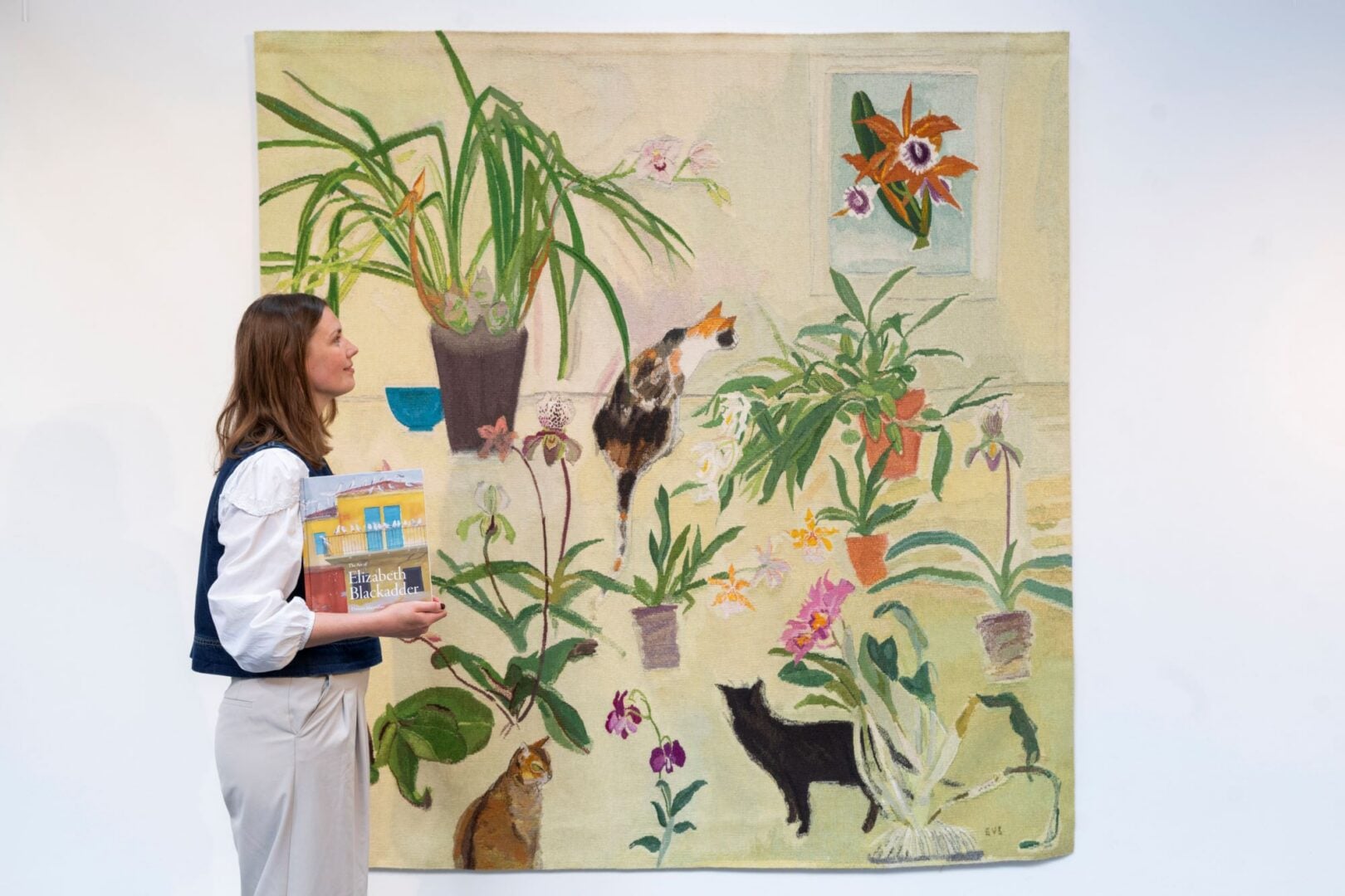 A woman looking at a painting on a wall, which features 3 cats and several plants.