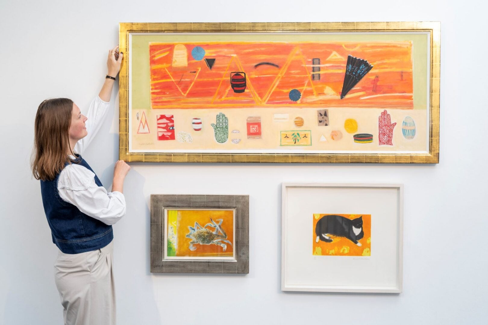 A woman standing at a wall, on which are displayed 3 colourful paintings.