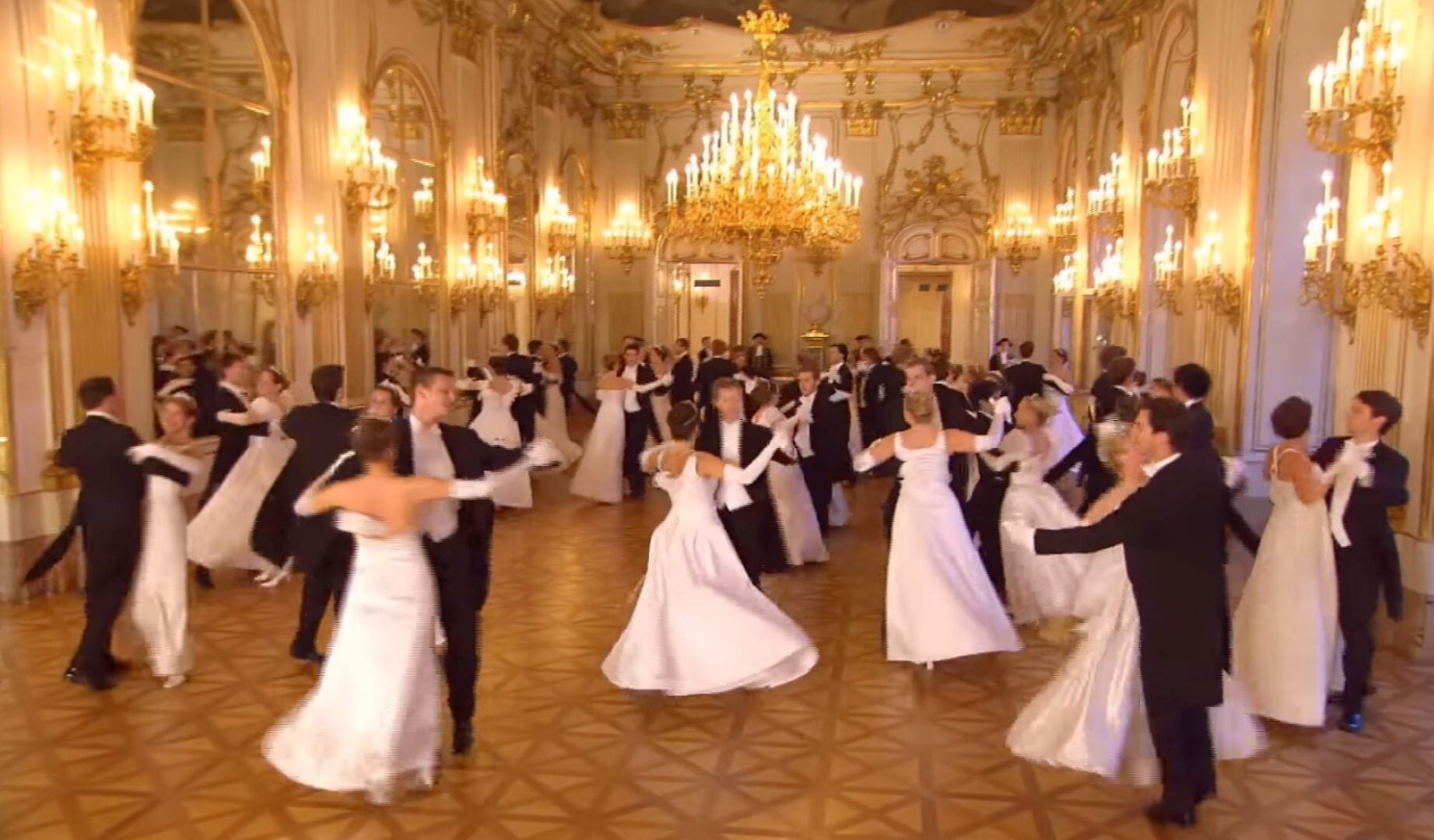 Couples in formal dress dancing in an opulent room, filled with chandeliers.