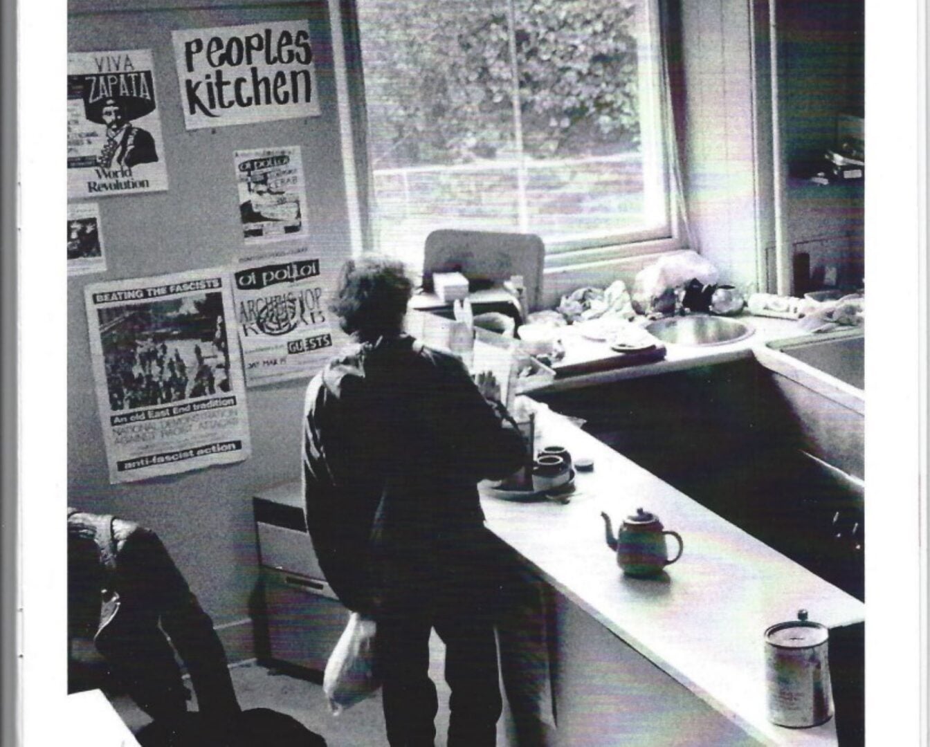 Black and white image of a person standing in a kitchen, with posters on the walls.