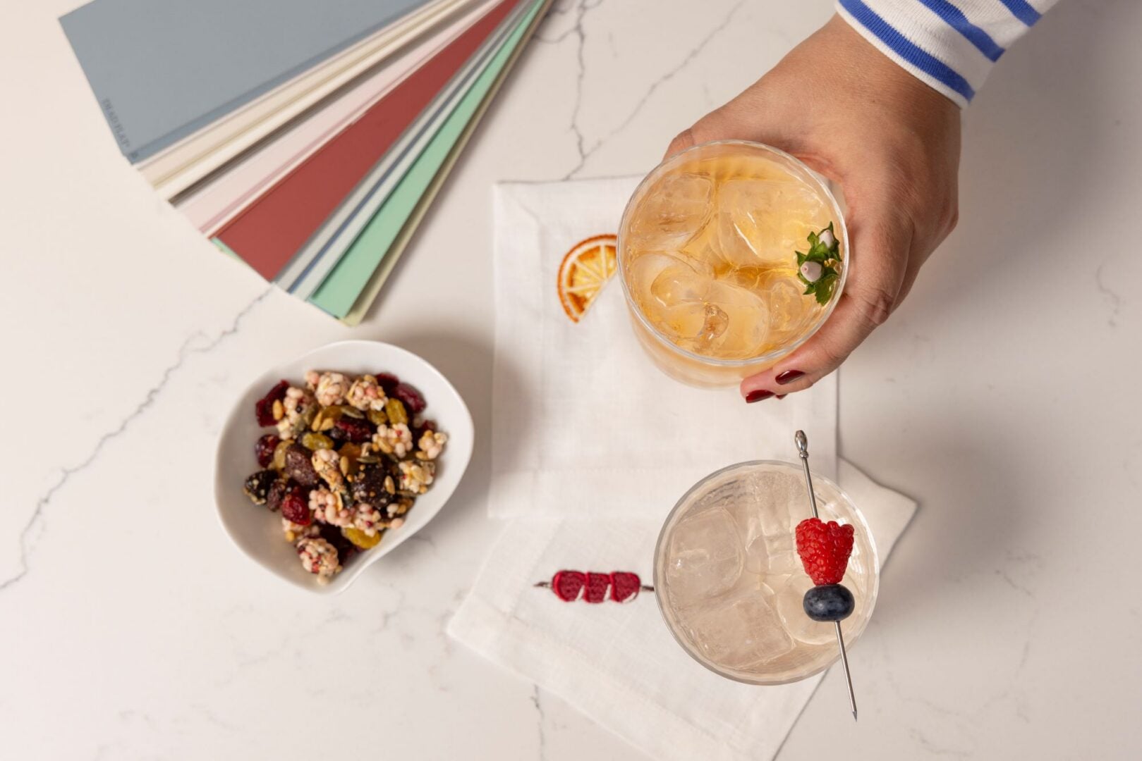 A hand placing a drink on a table, which also has on it a bowl of nuts and fruit pieces, and different coloured pieces of card.