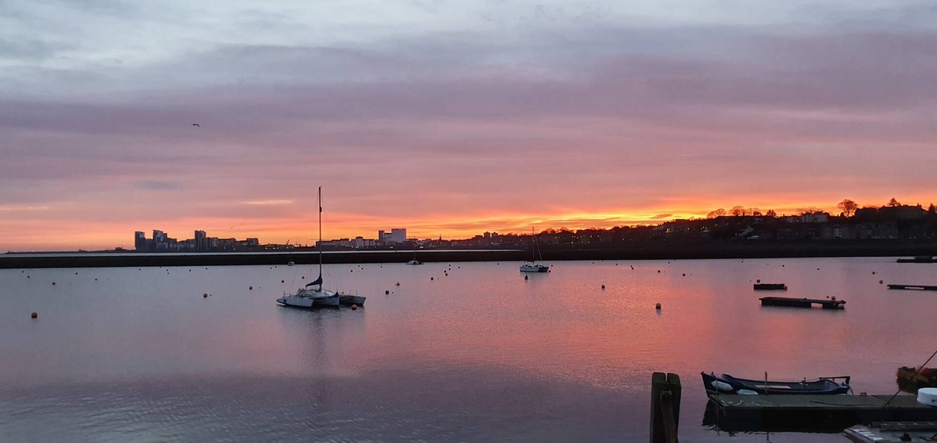 Sailboats moored on calm water at sunrise, with Edinburgh’s waterfront silhouetted beneath a pink and orange sky.,© Ross McDowell
