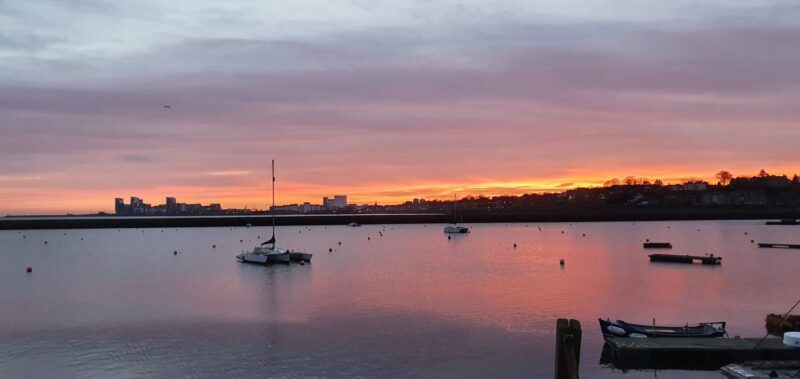 Sailboats moored on calm water at sunrise, with Edinburgh’s waterfront silhouetted beneath a pink and orange sky.,© Ross McDowell