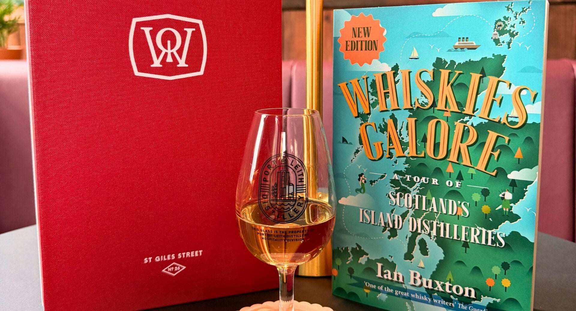 A book, titled Whiskies Galore! sitting on a table, with a glass of whisky in front of it.