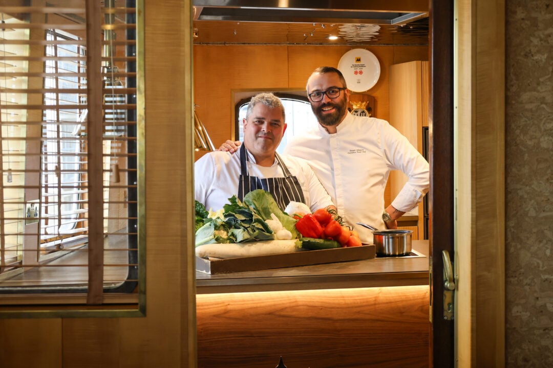 2 men dressed in chef's whites, standing in front of a box of vegetables.
