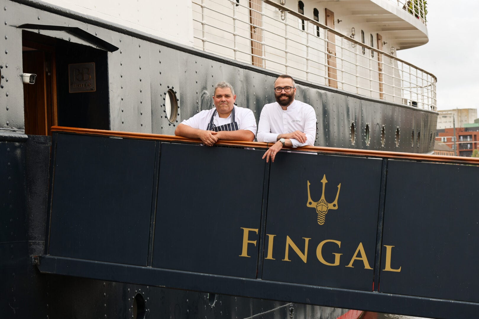 2 men dressed in chef's whites, standing on the gangway of a ship.