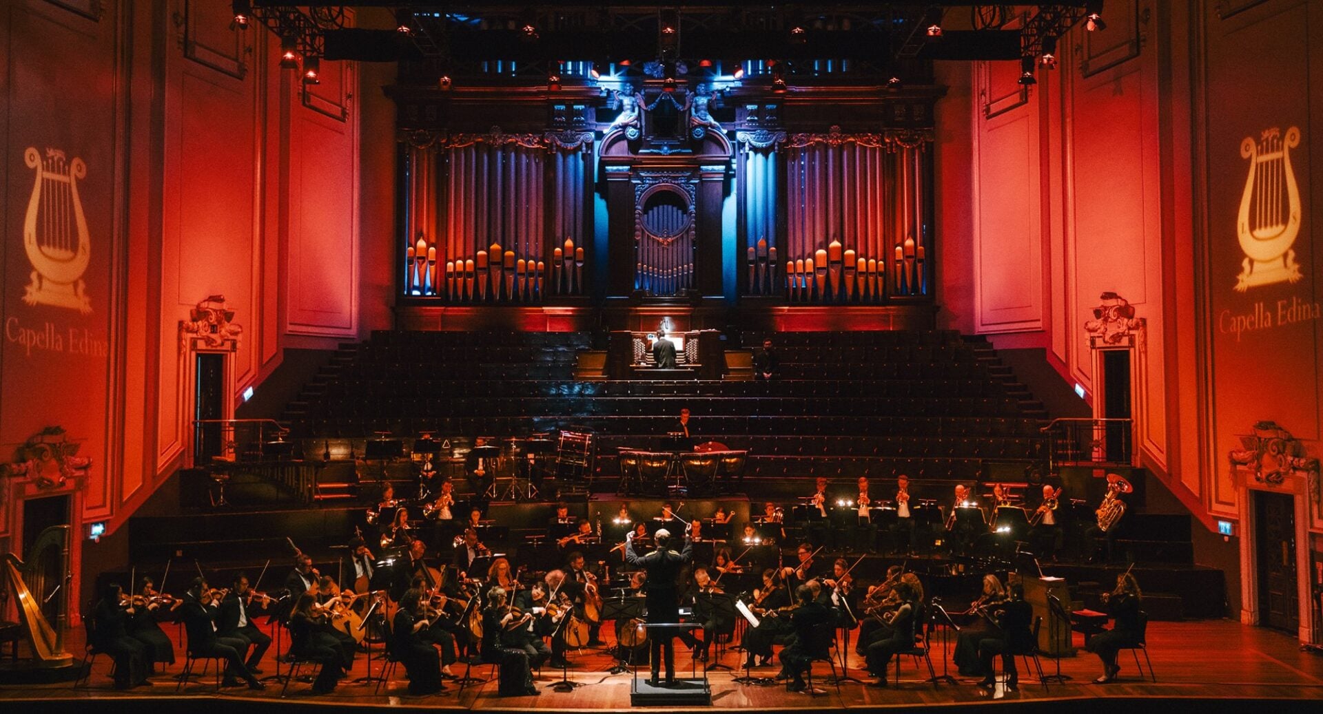 A full orchestra performing on stage, with an ornate grand organ in the background.