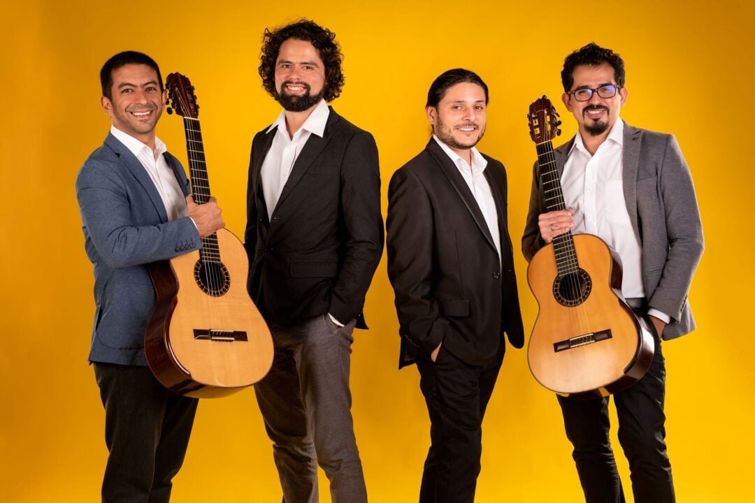 4 men standing in a row, with the 2 at each end each holding a guitar.