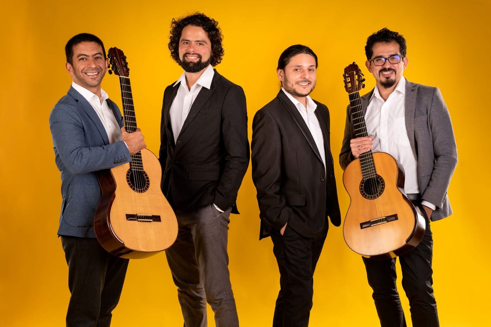4 men standing in a row, with the 2 at each end each holding a guitar.
