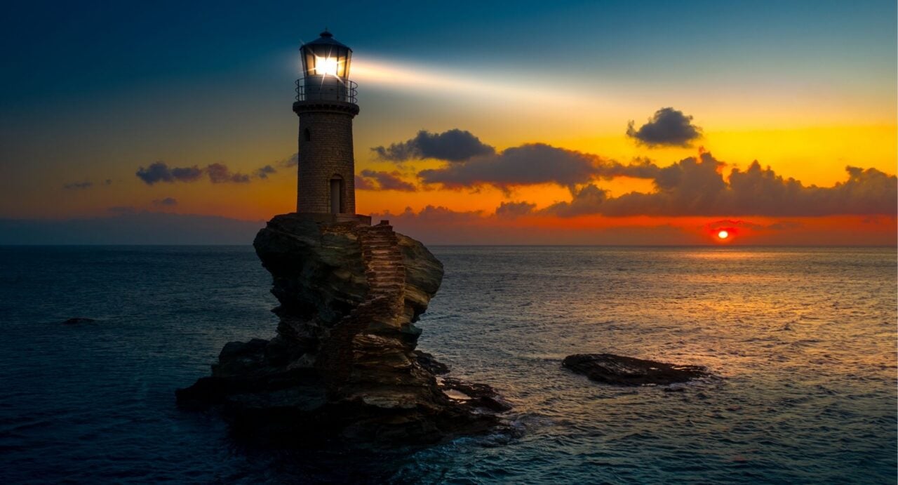 A lit lighthouse on a outcrop of rocks in the middle of an expanse of water.