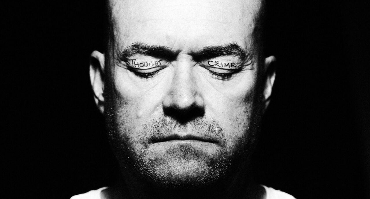 A black and white image of a man's face, with his eyes closed. On one eyelid is written 