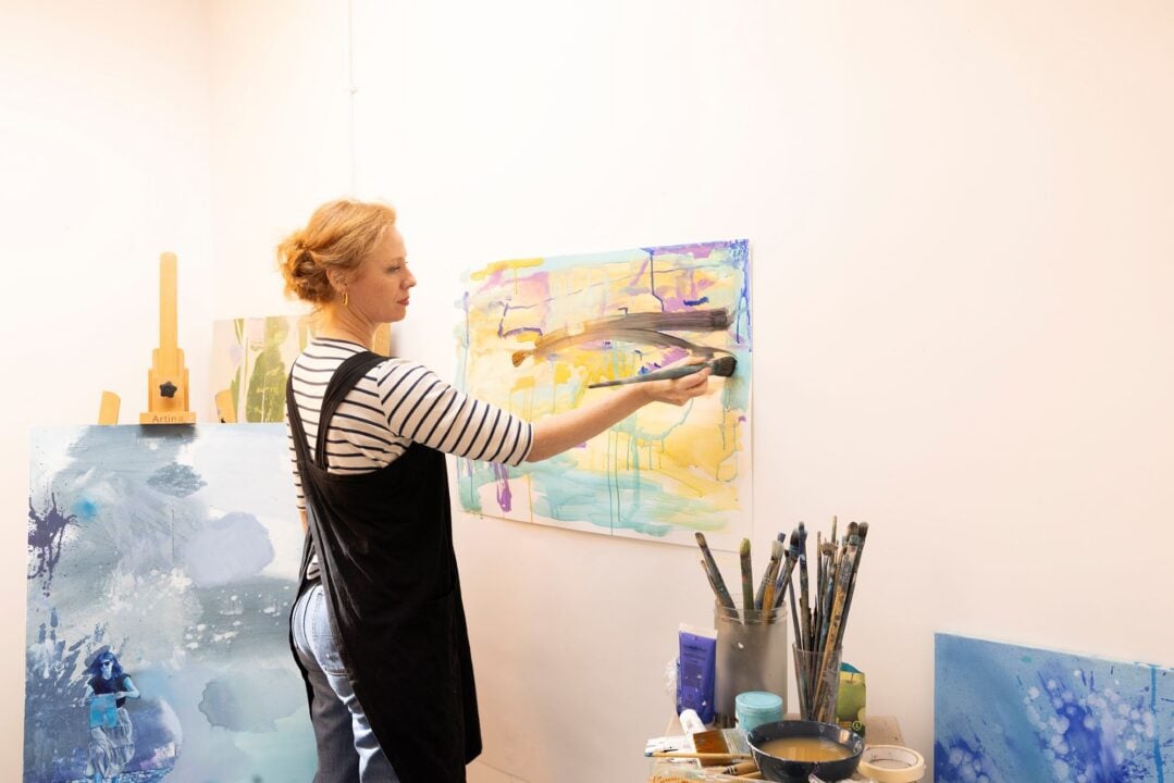 A woman standing in an arts studio, surrounded by paintings and brushes.