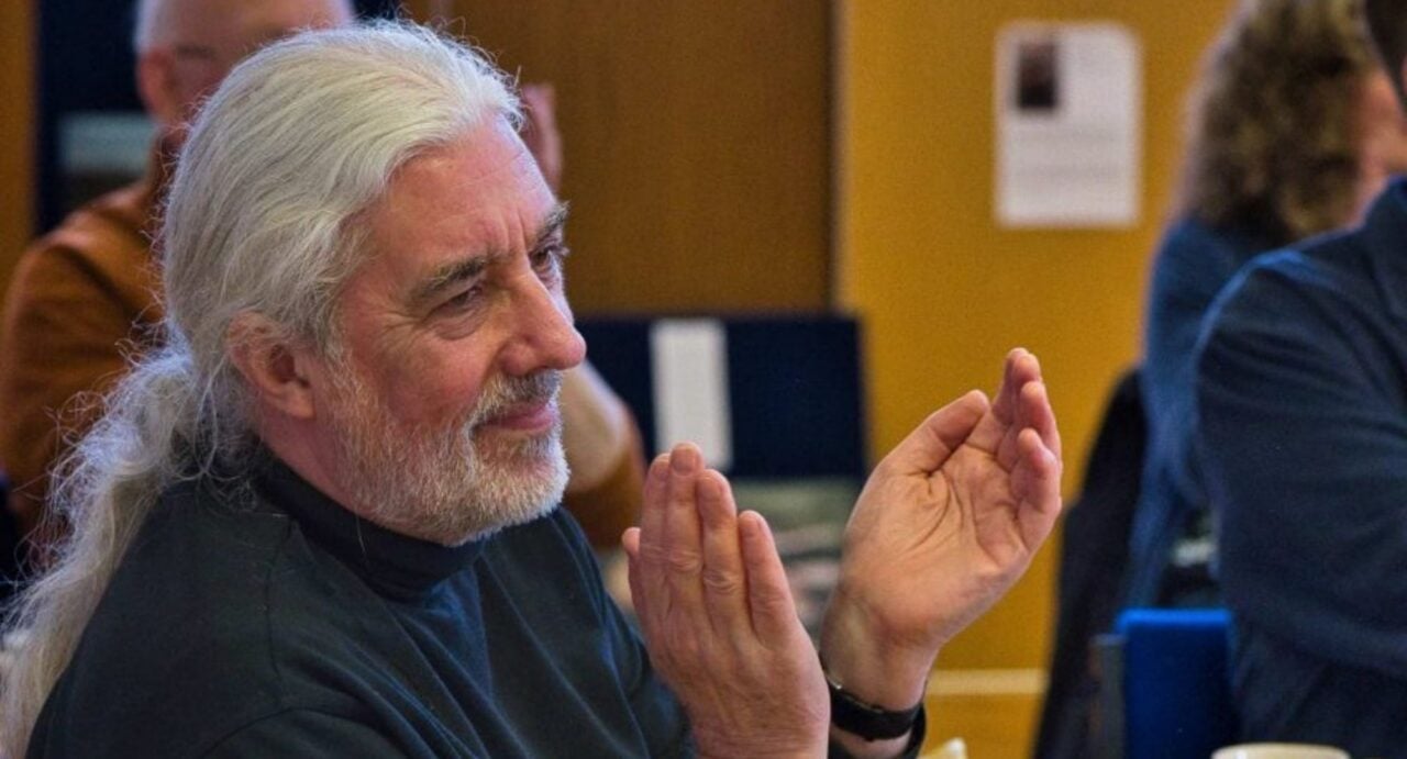 A man with long white hair and beard, clapping his hands.